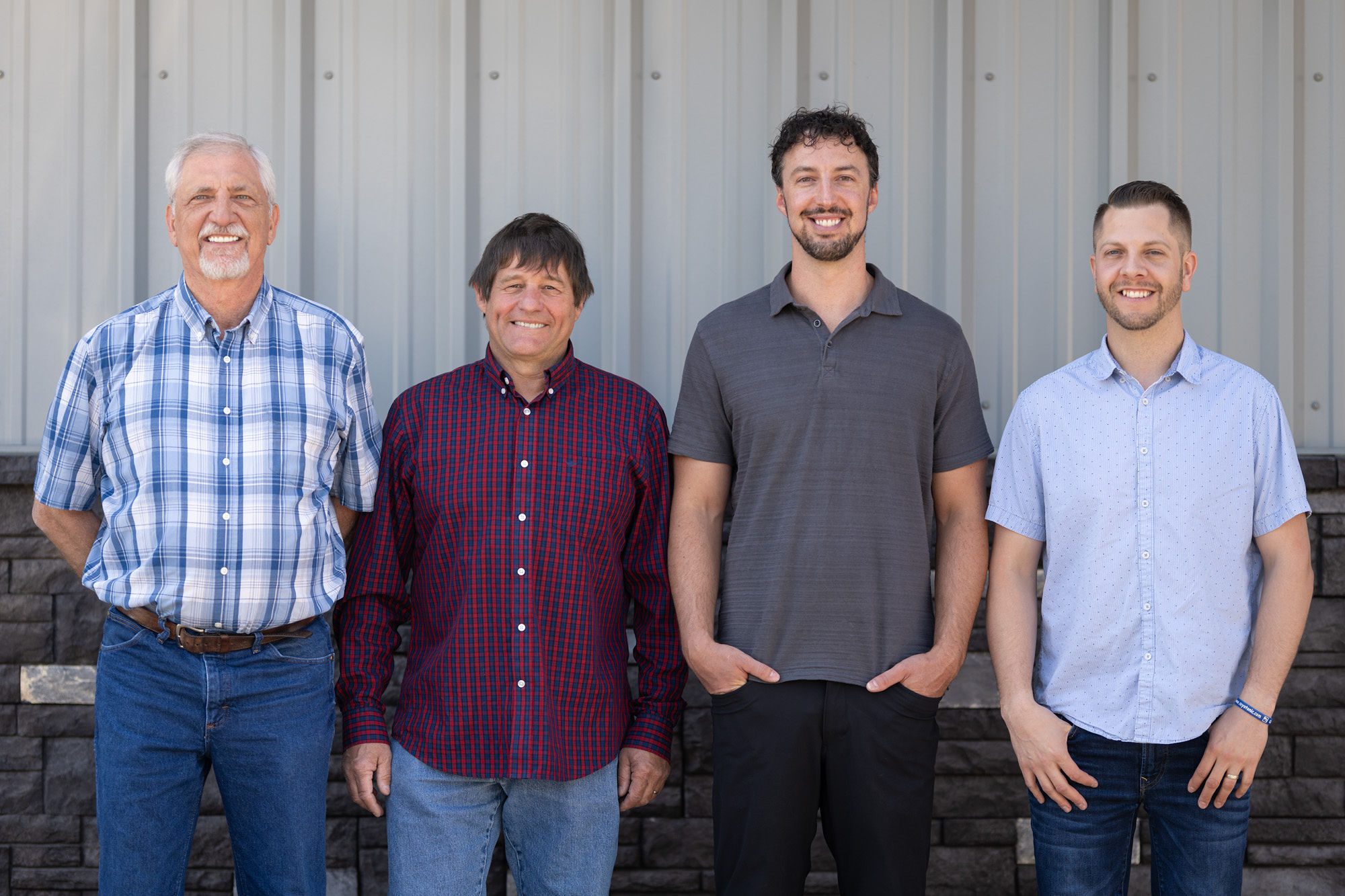 Four men stand in a row, smiling in front of a corrugated metal and brick wall. Each wears casual attire: two in button-up shirts, one in a polo, and one in a plaid shirt.