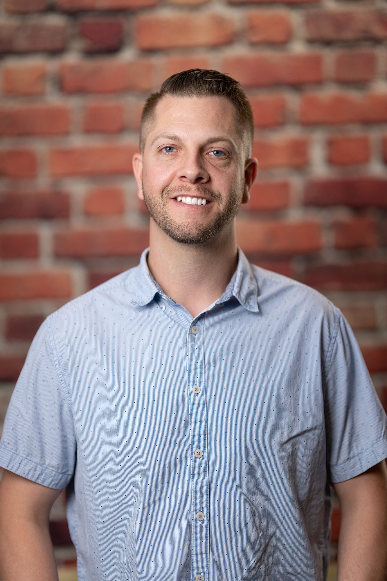 A man with short hair and a trimmed beard wearing a light blue, short-sleeved button-up shirt stands against a background of red brick.