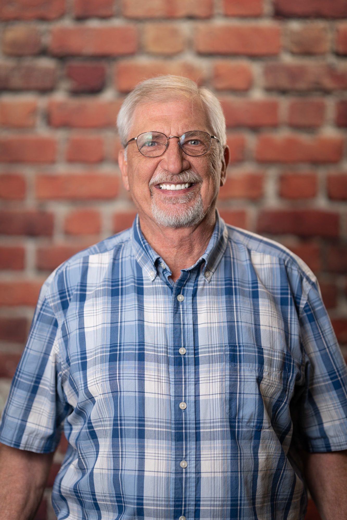 Elderly man with glasses and a short white beard smiling, wearing a blue plaid shirt, and standing in front of a brick wall.