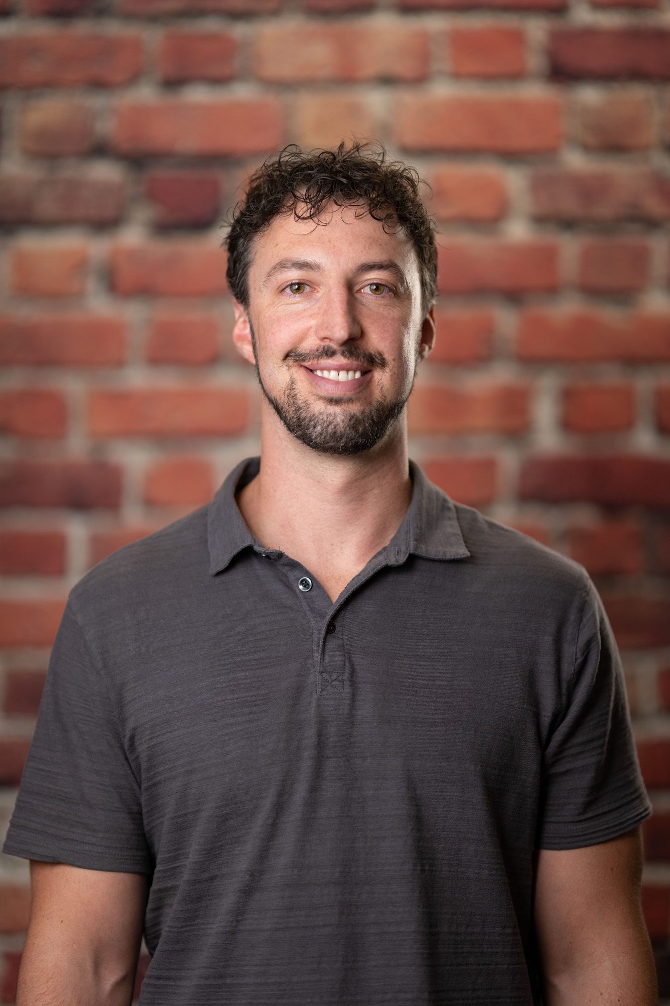 A man with short curly hair and a beard is smiling and wearing a gray polo shirt, standing in front of a brick wall.